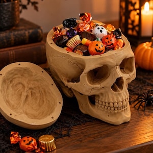 Pode incluir: Um recipiente em forma de crânio humano bege com tampa removível, cheio de vários doces de Halloween embrulhados, incluindo guloseimas em forma de abóbora, chocolates e doces com tema de fantasma, sobre uma superfície escura.