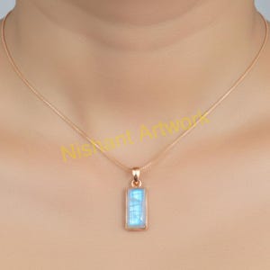 May include: A rose gold necklace featuring a rectangular pendant with a light blue gemstone. The pendant is suspended from a delicate chain. The gemstone has a subtle, iridescent quality, reflecting light. The necklace is worn around the neck.