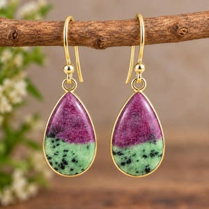 Può includere: Un paio di orecchini pendenti con pietre di rubino zoisite a forma di goccia incastonate in metallo dorato. Ogni pietra presenta una netta separazione di colore tra il magenta intenso e il verde chiaro maculato, appese a semplici ganci dorati.