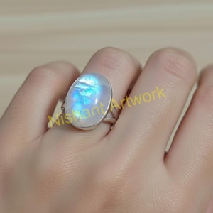 May include: Oval-shaped moonstone ring with a silver band. The moonstone has a pearly white base with blue and iridescent flashes. The ring is set on a hand, showcasing the gemstone's natural beauty and design.