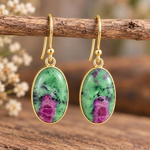 Può includere: Un paio di orecchini pendenti color oro con cabochon ovali in zoisite verde con inclusioni di rubino magenta. Ogni pietra è incastonata in una lunetta di metallo color oro liscio e pende da un semplice gancio per orecchie color oro.