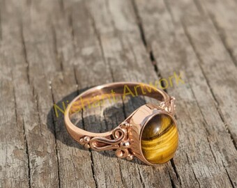 May include: A rose gold ring with a large, oval tiger's eye stone. The ring has an ornate, filigree design on the band and around the stone setting. The tiger's eye stone is a warm brown color with golden stripes.