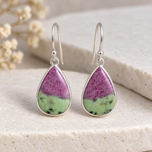 May include: A pair of drop earrings featuring teardrop-shaped stones with a dual-tone design of vibrant magenta and light green, accented with small black flecks. The stones are set in polished silver-toned metal frames with matching silver-toned hooks.