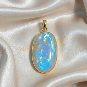 Puede incluir: Colgante ovalado con marco y asa dorados. El colgante presenta una gran piedra iridiscente azul y blanca. El fondo es una tela crema similar al satén. Se trata de una joya.