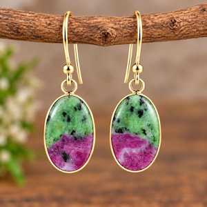May include: A pair of gold-toned drop earrings featuring oval-shaped cabochons of green and magenta zoisite with black inclusions. The stones are set in smooth gold-coloured metal bezels and hang from simple gold-toned shepherd hook ear wires.