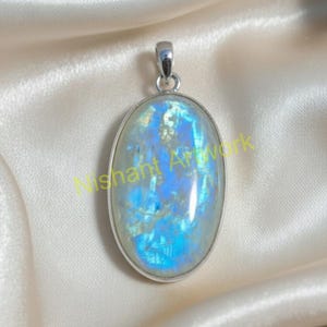 Puede incluir: Colgante ovalado con una gran piedra lunar iridiscente. La piedra tiene un color azul y blanco con un marco de metal plateado y una argolla. El colgante se muestra sobre una tela de satén.