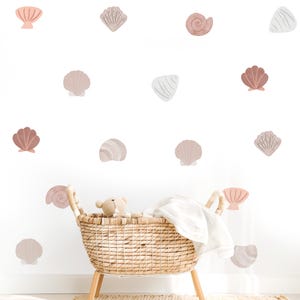 May include: A woven wicker basket with a light-colored teddy bear and a white cloth inside, positioned in front of a wall decorated with various seashell wall decals in shades of pink, beige, brown, and white, featuring diverse shell shapes.