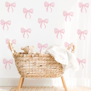 May include: A wicker basket with a white blanket inside, sitting in front of a white wall with pink watercolor bow decals.