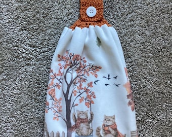 Peut inclure: Un essuie-main suspendu en tissu blanc imprimé de chats, de citrouilles et d'arbres. Le haut est doté d'une attache crochetée orange brûlé avec un bouton blanc à quatre trous. Le motif inclut des feuilles d'automne, des oiseaux et une clôture.