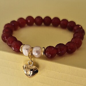 May include: A beaded bracelet featuring faceted, deep red beads and two white pearls. A gold-colored heart charm dangles from the bracelet. The bracelet is displayed on a yellow surface.