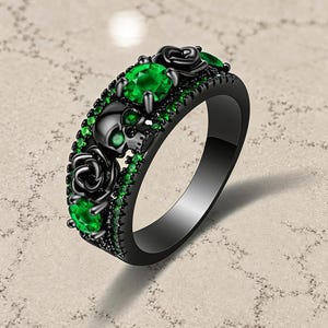 May include: A black metal ring featuring a central skull motif flanked by two sculpted black roses. The design is accented with multiple round, vibrant green gemstones set along the band and within the eye sockets of the skull figure.