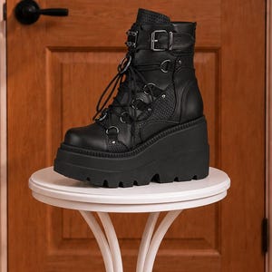 May include: A black ankle boot featuring a high platform wedge sole with a lugged tread. The upper is constructed from smooth and textured snake-patterned panels, accented with silver-toned metal hardware, D-ring eyelets, black laces, and a buckle.