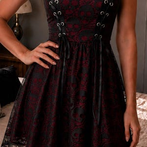 May include: A sleeveless mini dress featuring a black lace overlay with a skull pattern over a dark red base. The bodice includes two vertical black ribbon lace-up details with silver grommets, thin shoulder straps, and a scalloped lace hemline.