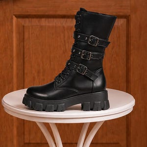 May include: A black mid-calf boot featuring a thick lug sole, a lace-up front, and three horizontal straps across the shaft, each secured with a silver-toned metal buckle. The boot is constructed with a smooth finish and a rounded toe design.