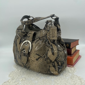 Puede incluir: Un bolso de mano marrón y negro con estampado de piel de serpiente y cierre de hebilla plateada. El bolso tiene un asa superior y una pequeña etiqueta adjunta. El bolso está sobre una superficie blanca.