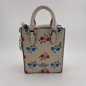 May include: A miniature cream-coloured handbag with a top handle and a floral pattern in red and blue. The bag features silver-tone hardware and a small charm. The brand name "COACH" is visible on the front.