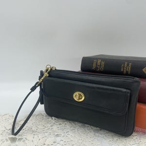 Puede incluir: Bolsa de muñeca de cuero negro con cierre y herrajes dorados. La bolsa de muñeca tiene un bolsillo frontal y una correa para la muñeca. Libros en el fondo.