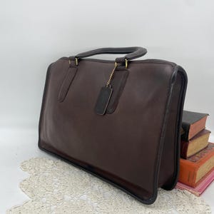 Può includere: Una ventiquattrore in pelle marrone con manico superiore e forma rettangolare. La borsa ha finiture nere e hardware color oro. Una piccola etichetta in pelle pende dal manico. La ventiquattrore è posizionata su una superficie bianca.