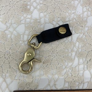 May include: A black leather strap with a gold-toned metal snap button attached to a brass swivel lobster clasp. The item is positioned above a wooden ruler that shows the total length of the object is approximately 4 inches in total length.