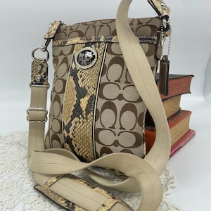 Może przedstawiać: Brązowa torba na ramię Coach z paskiem ze skóry węża pośrodku. Torba posiada beżowy pasek i logo Coach. Torba leży na białej koronkowej powierzchni.