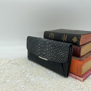 May include: Black wallet with a textured, snakeskin-like flap and a silver-tone accent. The wallet is rectangular and closed. The background includes vintage books and a cream-colored lace doily.