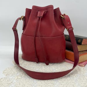 May include: A red leather bucket bag with a drawstring closure and a long shoulder strap. The bag has a front pocket and gold-tone hardware. The bag is approximately 10 inches tall.