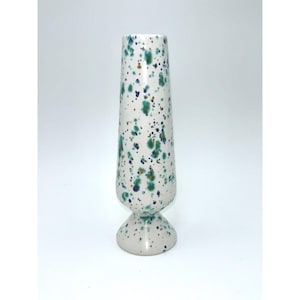 May include: A tall, slender ceramic vase with a flared base, featuring a glossy off-white finish decorated with a speckled pattern of dark blue, forest green, and metallic gold paint splatters distributed across the entire exterior surface.