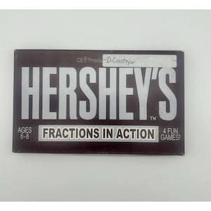 Könnte beinhalten: Eine rechteckige Hershey's Fractions in Action Spieleschachtel mit dunkelbraunem Hintergrund und silberner Schrift. Die Schachtel zeigt den Text Ages 6-8, 4 Fun Games und einen handschriftlichen Namen D. Gasteyer auf einem weißen Etikett oben.