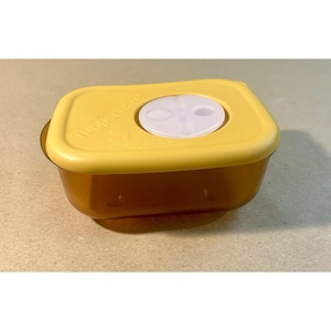 Puede incluir: Un recipiente Tupperware marrón y amarillo. El recipiente tiene forma rectangular con una tapa amarilla y una ventilación blanca. La tapa tiene el logotipo de Tupperware en relieve.
