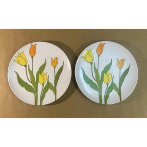 Op de afbeelding: Twee witte keramische borden met een tulpenontwerp. Elk bord heeft gele en oranje tulpenbloemen met groene stelen en bladeren. De borden zijn rond en hebben een eenvoudig, elegant ontwerp.