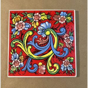 May include: Square ceramic tile with a vibrant floral design. The tile features a red background with colorful flowers in shades of pink, blue, yellow, and green. The floral pattern is symmetrical and detailed, with a white border.