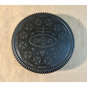 Op de afbeelding: Een zwarte Oreo-koekje met een gedetailleerd reliëfontwerp. Het koekje heeft het woord "OREO" in het midden, omringd door geometrische patronen en een geribbelde rand. Het koekje staat tegen een neutrale achtergrond.