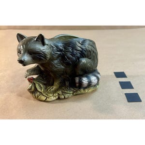May include: A detailed figurine of a raccoon, painted in shades of grey and black, with a white and black striped tail. The raccoon is perched on a bed of green leaves with red berries. The figurine is likely ceramic or a similar material.