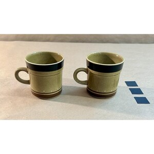 May include: Two ceramic mugs with a green-brown base color, black rim, and brown and white stripes. Each mug has a curved handle. The mugs are on a neutral surface with three small blue squares.