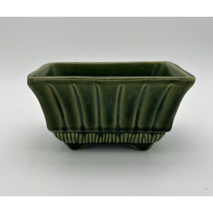May include: A rectangular ceramic planter with a deep olive green glaze. The exterior features vertical fluted ridges along the sides and a textured, ribbed pattern near the base. The vessel is supported by four small, integrated feet at the corners.