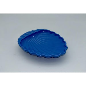 May include: A bright blue dish shaped like a seashell with a scalloped edge and a smooth, glossy finish. The interior of the dish features raised, radiating ridges that mimic the natural texture of a clam shell, creating a concave surface.