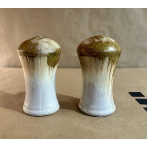 May include: Two ceramic salt and pepper shakers. The shakers are white with a brown and green top. The glaze drips down the sides of the shakers. The shakers have a rounded top and a flared base.