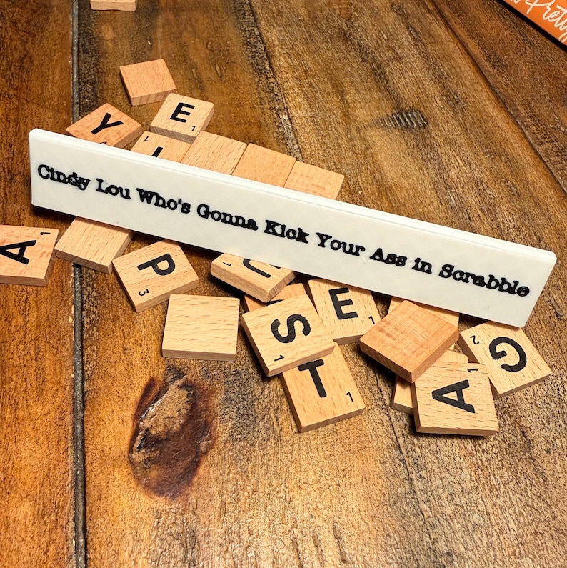Puede incluir: Un soporte blanco para fichas de Scrabble con el texto "Cindy Lou Who's Gonna Kick Your Ass in Scrabble" en letras negras. Fichas de Scrabble de madera con letras y n&uacute;meros negros est&aacute;n dispersas. Las piezas del juego est&aacute;n sobre una superficie de madera.