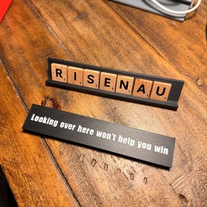 Puede incluir: Dos soportes negros con texto blanco sobre una superficie de madera. El soporte superior muestra la palabra "RISENAU" con letras de madera. El soporte inferior tiene el texto "Looking over here won't help you win."