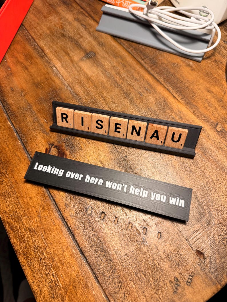 Puede incluir: Dos soportes negros con texto blanco sobre una superficie de madera. El soporte superior muestra la palabra "RISENAU" con letras de madera. El soporte inferior tiene el texto "Looking over here won't help you win."