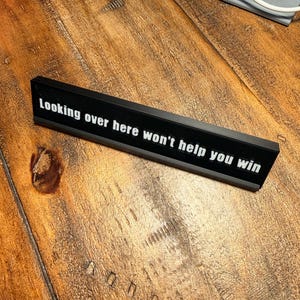 Puede incluir: Un letrero de escritorio rectangular negro con el texto "Looking over here won't help you win" en blanco. El letrero est&aacute; sobre una superficie de madera.