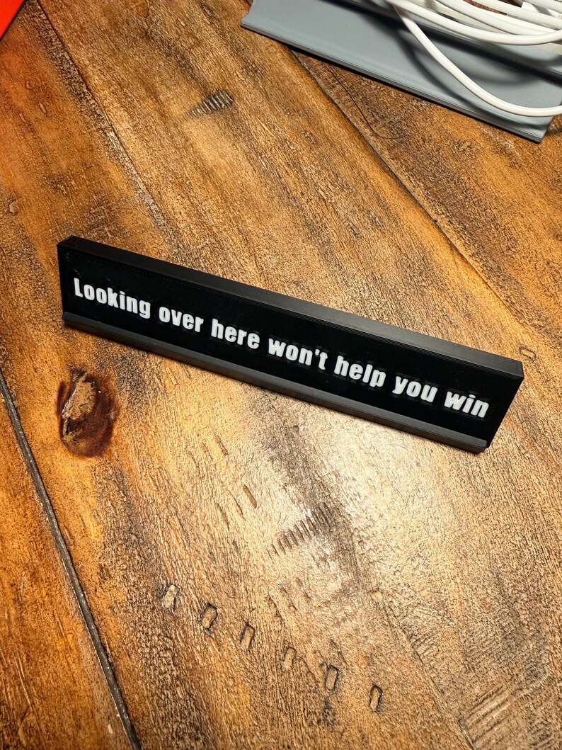 Puede incluir: Un letrero de escritorio rectangular negro con el texto "Looking over here won't help you win" en blanco. El letrero est&aacute; sobre una superficie de madera.