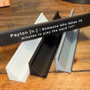 Puede incluir: Cuatro piezas decorativas rectangulares anguladas en blanco, negro y gris, con una pieza negra encima. La pieza negra tiene texto blanco que dice: "Payton [n.]: Alguien que tarda 10 minutos en jugar la palabra 'IT'".