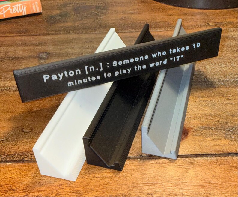 Puede incluir: Cuatro piezas decorativas rectangulares anguladas en blanco, negro y gris, con una pieza negra encima. La pieza negra tiene texto blanco que dice: "Payton [n.]: Alguien que tarda 10 minutos en jugar la palabra 'IT'".