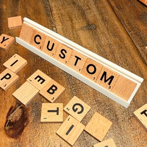 Puede incluir: Fichas de Scrabble de madera que deletrean "CUSTOM" en un estante de fichas blanco, con fichas adicionales esparcidas alrededor. Las fichas tienen letras y n&uacute;meros negros. El fondo es una superficie de madera.
