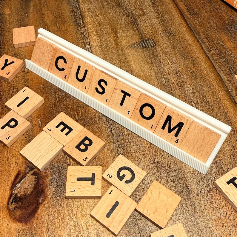 Puede incluir: Fichas de Scrabble de madera que deletrean "CUSTOM" en un estante de fichas blanco, con fichas adicionales esparcidas alrededor. Las fichas tienen letras y n&uacute;meros negros. El fondo es una superficie de madera.