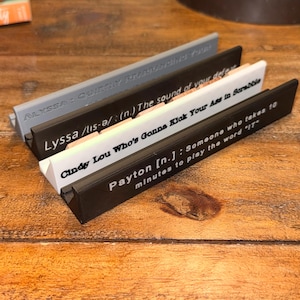 Puede incluir: Cuatro soportes para fichas de Scrabble en gris, negro y blanco, cada uno con una cita diferente impresa. Las citas incluyen "ALYSSA: QUIETLY REARRANGING YOUR", "Lyssa /lis-ə/ (n.) The sound of your defeat", "Cindy Lou Who's Gonna Kick Your Ass in Scrabble" y "Payton [n.]: Someone who takes 10 minutes to play the word 'IT'".