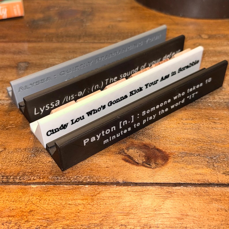 Puede incluir: Cuatro soportes para fichas de Scrabble en gris, negro y blanco, cada uno con una cita diferente impresa. Las citas incluyen "ALYSSA: QUIETLY REARRANGING YOUR", "Lyssa /lis-ə/ (n.) The sound of your defeat", "Cindy Lou Who's Gonna Kick Your Ass in Scrabble" y "Payton [n.]: Someone who takes 10 minutes to play the word 'IT'".