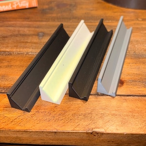 Puede incluir: Cuatro rieles angulares para marcos de fotos en negro, blanco, negro y gris. Los rieles est&aacute;n dispuestos diagonalmente sobre una superficie de madera. Los marcos tienen forma triangular y est&aacute;n dise&ntilde;ados para contener una foto o una obra de arte.