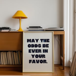 May include: A framed print with the text "MAY THE ODDS BE EVER IN YOUR FAVOR" in navy blue block letters. The print is displayed in a room with a yellow lamp, a wooden cabinet, and a turntable.